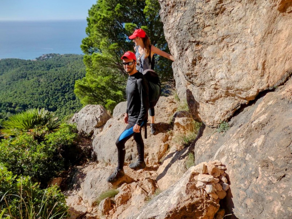 Dos excursionistas avanzan por un sendero rocoso en el  Pas de sa Trapa, con vistas al mar y vegetación abundante alrededor.