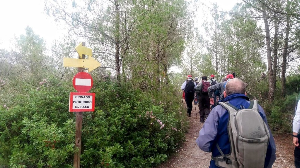 Grupo de personas caminando por un sendero rodeado de árboles y plantas, con un letrero que indica 'Privado Prohibido el Paso' y una flecha amarilla que señala la dirección.
