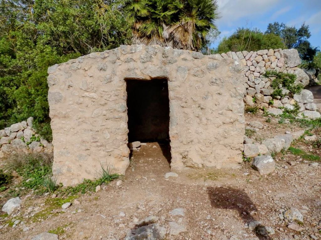 Edificio de piedra con una entrada abierta, rodeado de vegetación y piedras sueltas, ubicado en un área montañosa.