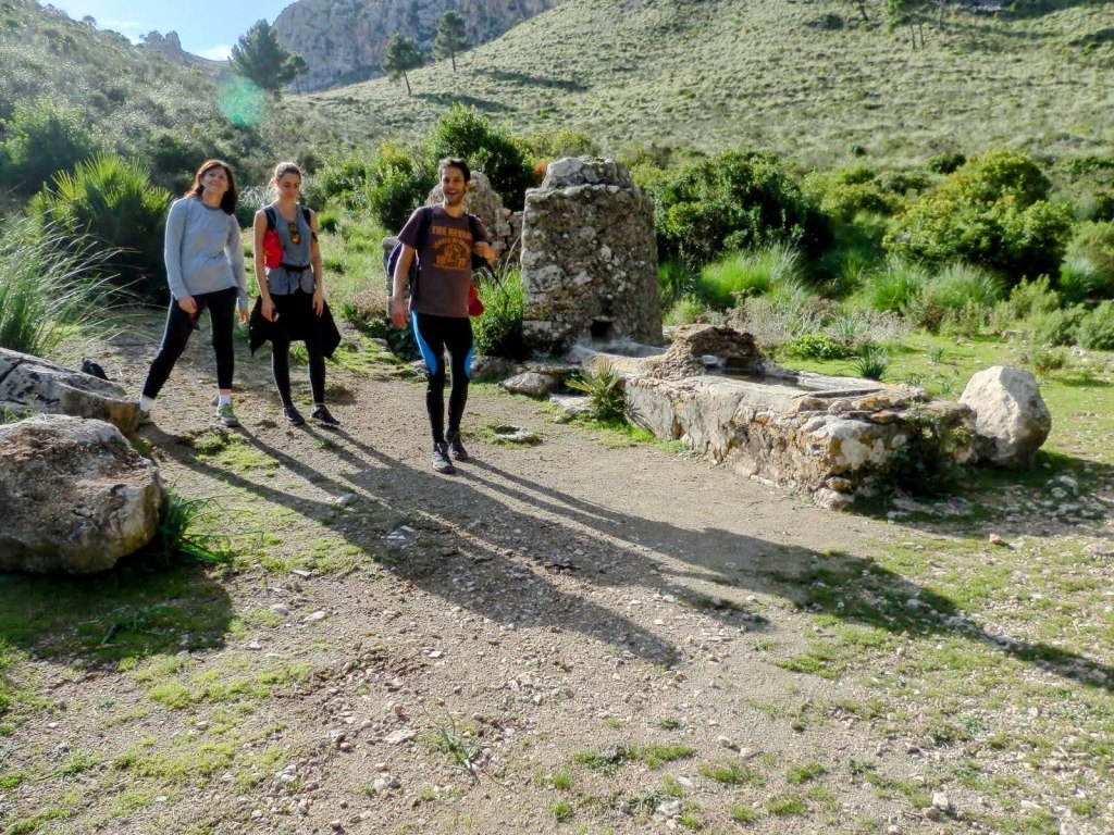 Grupo de tres personas caminando por un sendero en un paisaje montañoso, con vegetación y el Pozo de Ses Sinies al fondo.