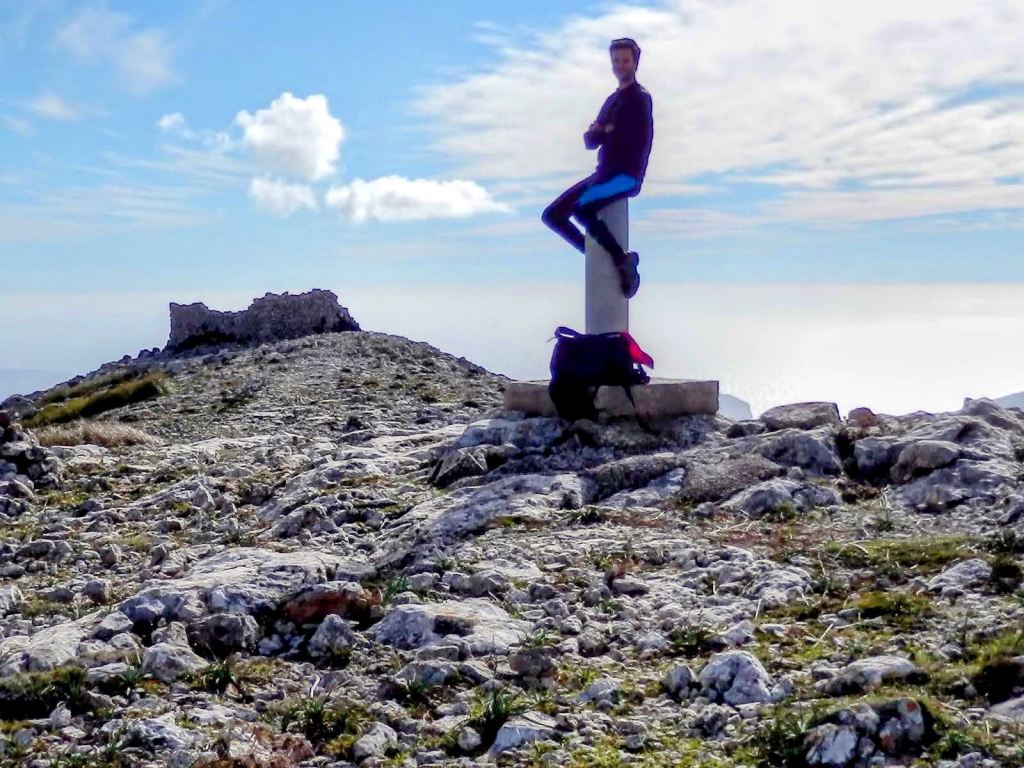 Persona descansando en la cima de la Mola de s'Esclop, con un vértice geodésico y ruinas de una construcción al fondo, rodeado de un paisaje montañoso bajo un cielo nublado.