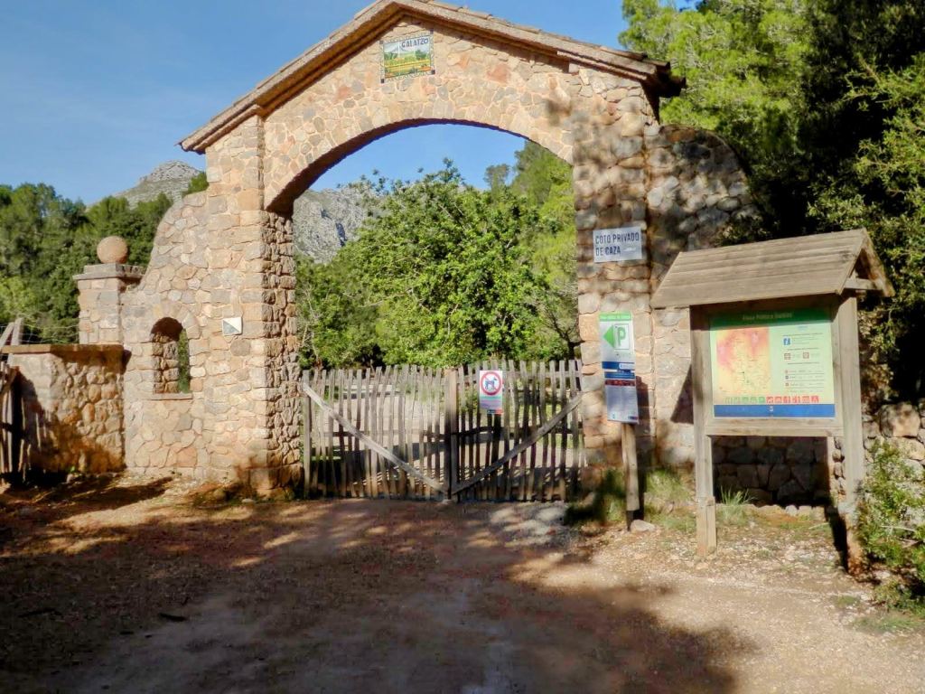 Entrada a la Finca Pública de Galatzó, mostrando un arco de piedra, una puerta de madera cerrada y un panel informativo junto a un camino.