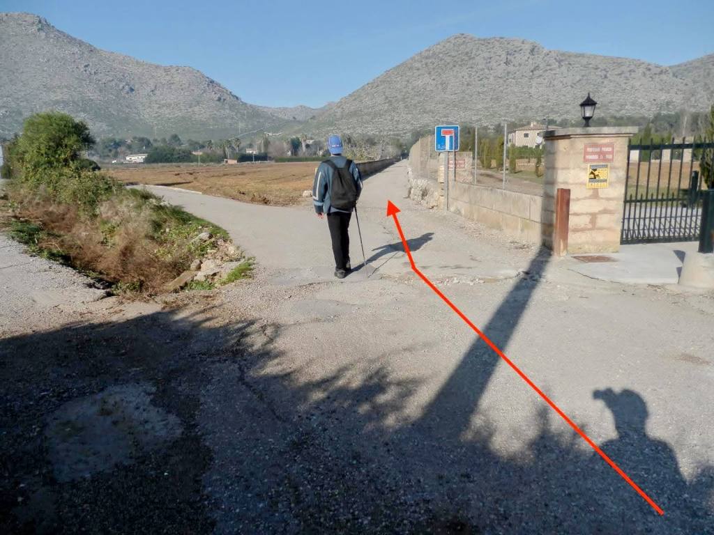 Persona caminando por un camino de grava junto a un portón de entrada, con montañas al fondo.