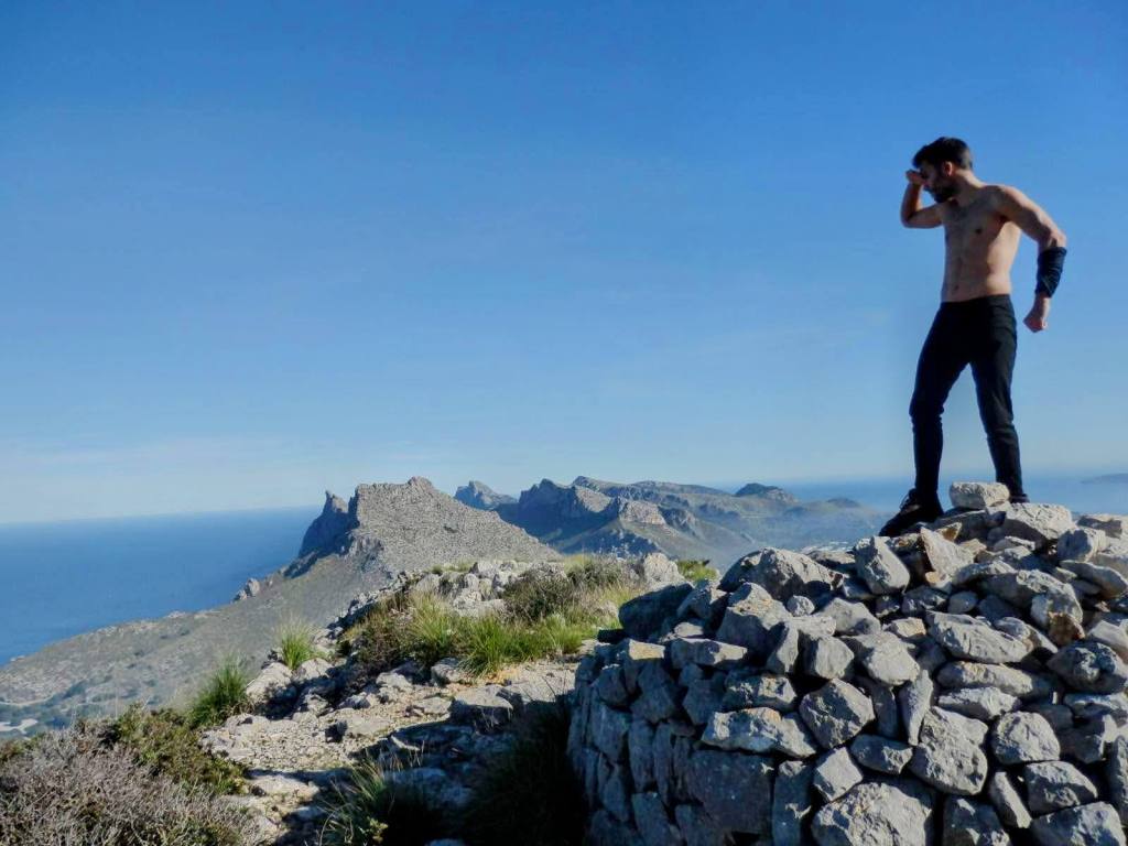 Un hombre de pie sobre un montón de piedras en la cima del Puig del Vilar, con vistas panorámicas al mar y montañas en el fondo.