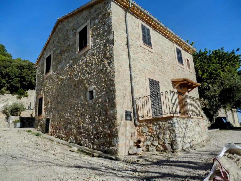 Vista de una casa de piedra con ventanas y un balcón, situada en Biniarroi, en un entorno natural con vegetación alrededor.