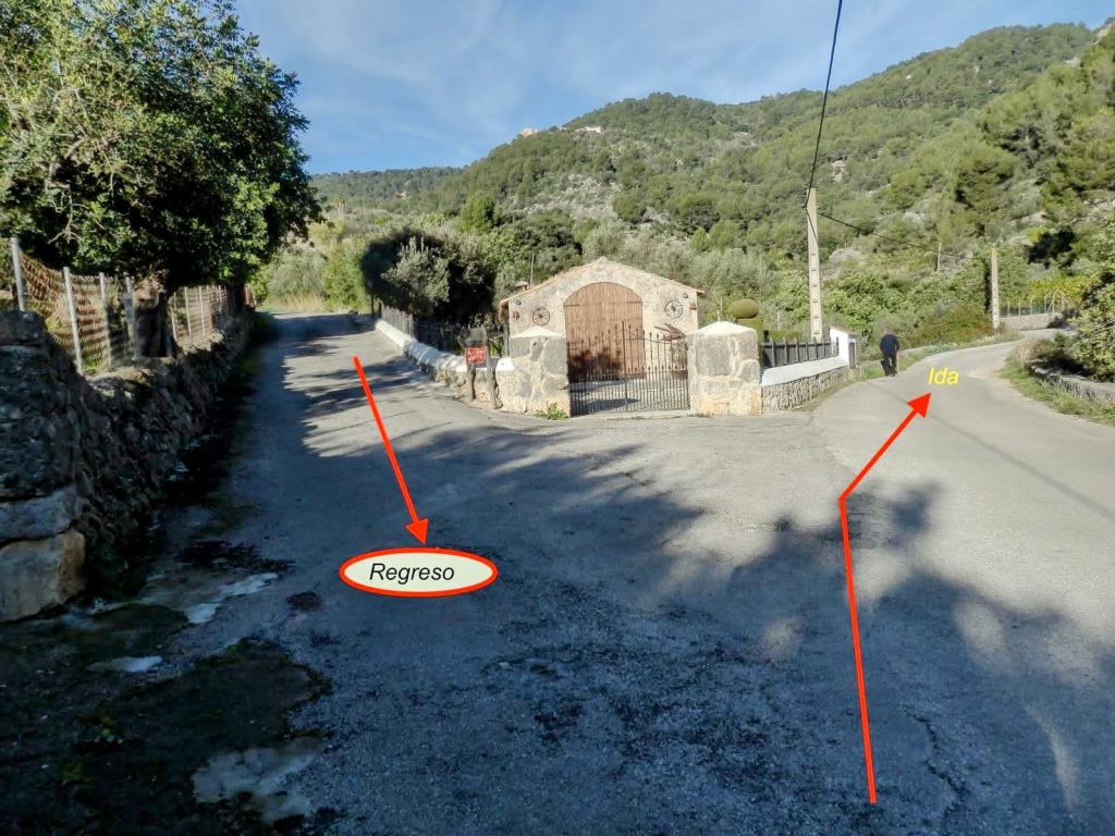 Señalización de bifurcación en la ruta Biniarroi-Camí de ses Cases Noves, con indicaciones para el regreso e ida, rodeada de vegetación y montañas.