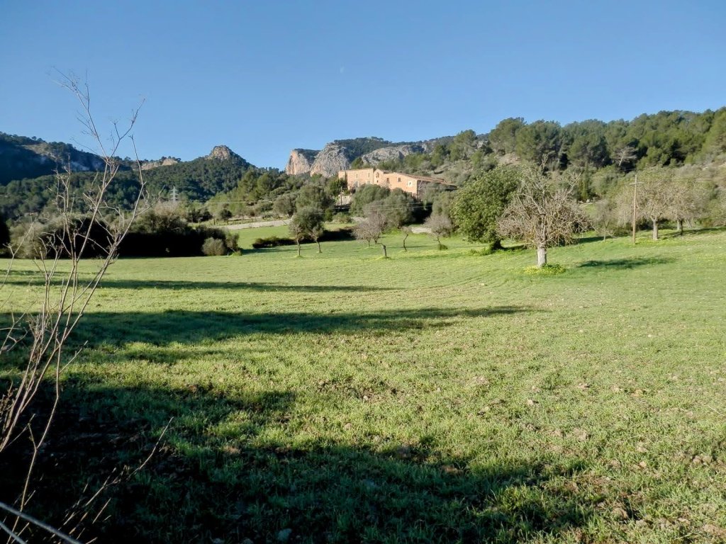 Panorama de un paisaje rural con un campo verde y colinas en el fondo, incluyendo una construcción histórica en la parte superior, bajo un cielo despejado.