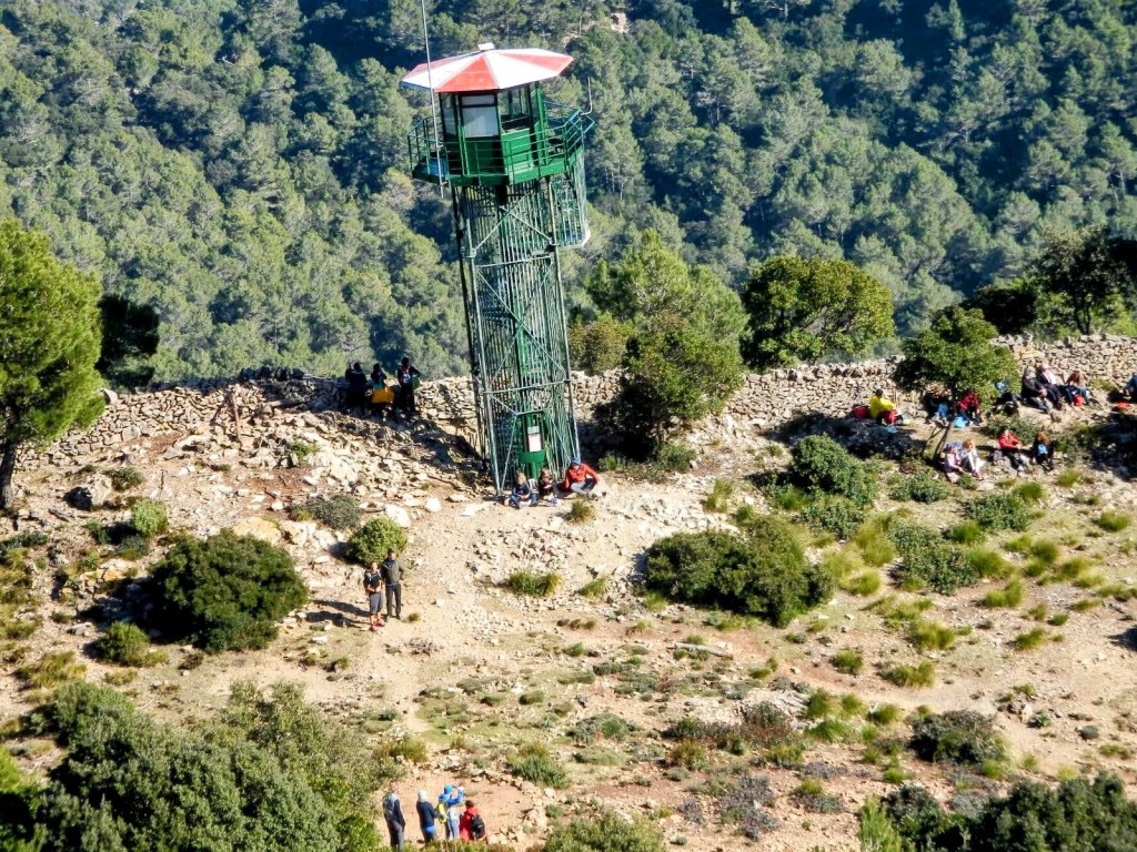 Vista panorámica de una torre de vigilancia contra incendios en un área montañosa con grupos de personas sentadas en el suelo y árboles alrededor.