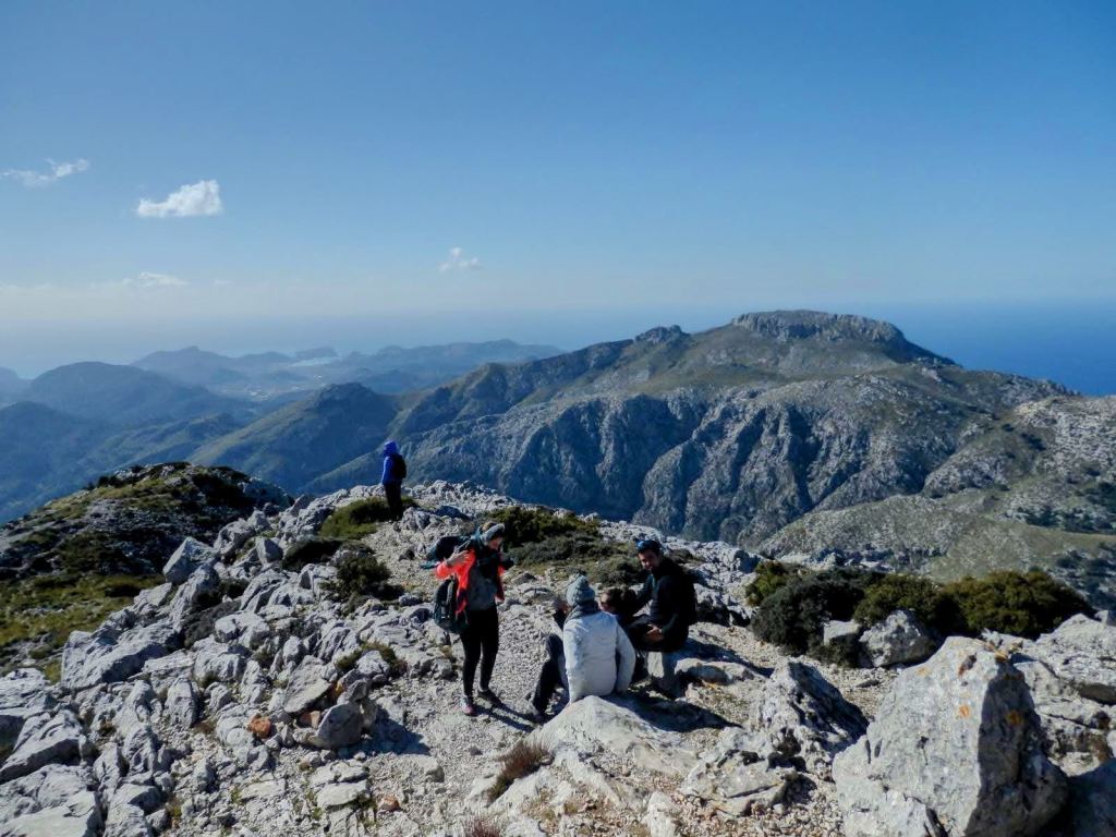 Vistas panorámicas desde la cima del Puig de Galatzó, con varias personas descansando en un área rocosa y un paisaje montañoso de fondo.