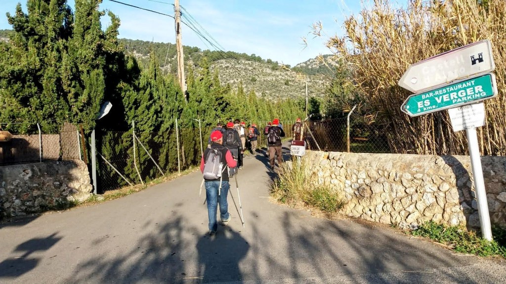 Grupo de senderistas caminando por el Camí de sa Sort (GR-221).con carteles indicando dirección al restaurante 'Es Verger' y al 'Castell d'Alaró'.
