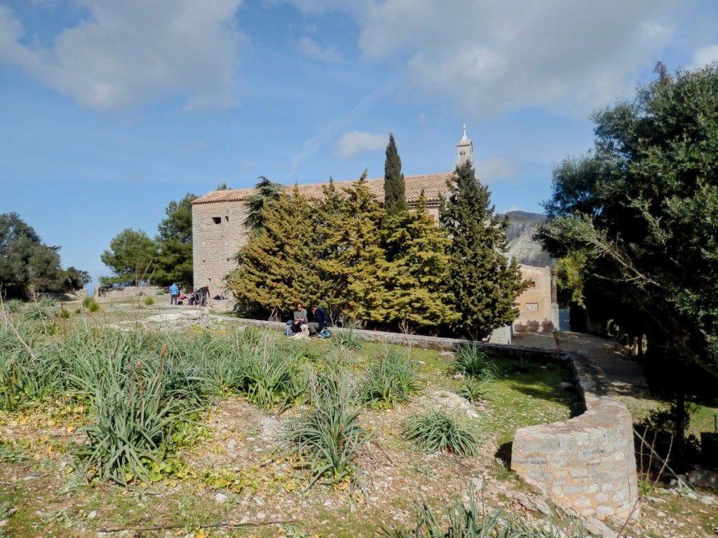 Vista exterior de la Ermita de Betlem rodeada de vegetación y personas en la entrada, con un cielo parcialmente nublado.