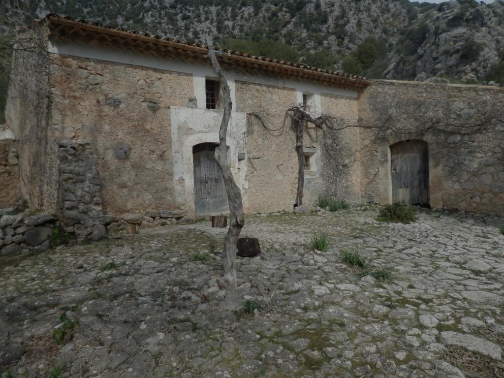 casas de ses Figueroles con paredes de piedra y techo de tejas, ubicada en un entorno montañoso, con un suelo empedrado y una vegetación escasa.