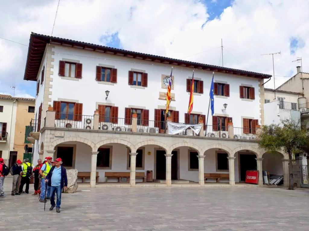 Edificio de dos plantas en Sant Llorenç de Cardessar, con un balcón y varias banderas en la fachada. Un grupo de personas se encuentra en la plaza frente al edificio.