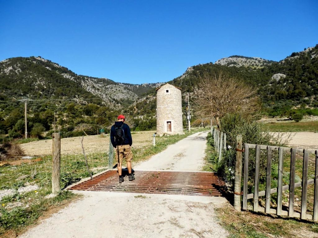 Un senderista con mochila se encuentra frente a una antigua torre de piedra en un camino rural rodeado de montañas y naturaleza, bajo un cielo despejado.