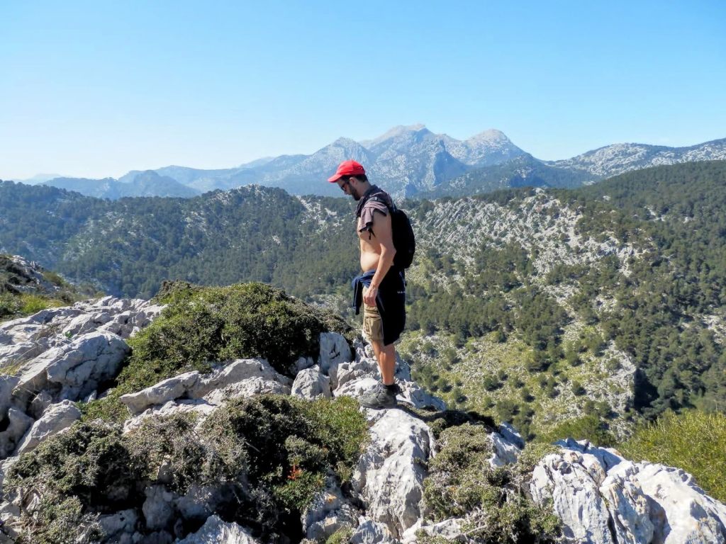 Hombre observando el paisaje desde la cima del Puig d’en Costurer, rodeado de rocas y vegetación, con vistas a montañas y valles al fondo.