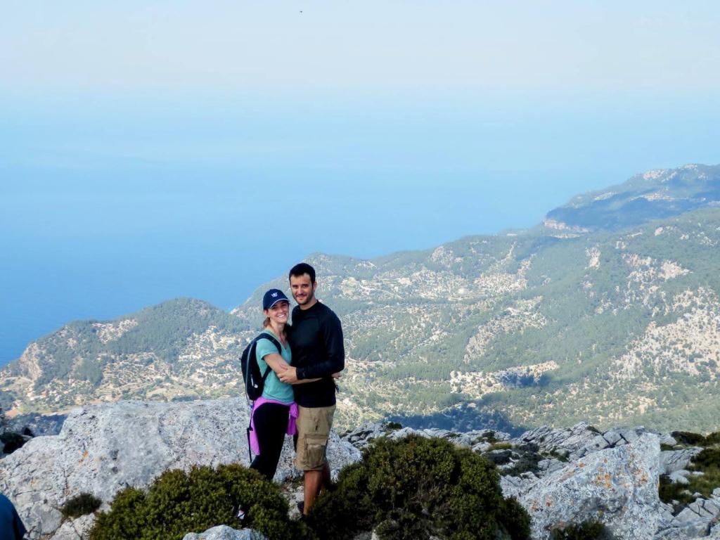 Pareja sonriendo y abrazándose en la cima del Puig de Galatzó, con vistas panorámicas de montañas y el mar de fondo.