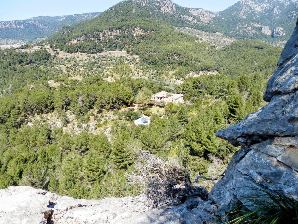 Vista panorámica desde una roca que muestra la  casa de Ses Esteles rodeada de árboles en un área montañosa, en la Ruta Puig de Na Fátima.