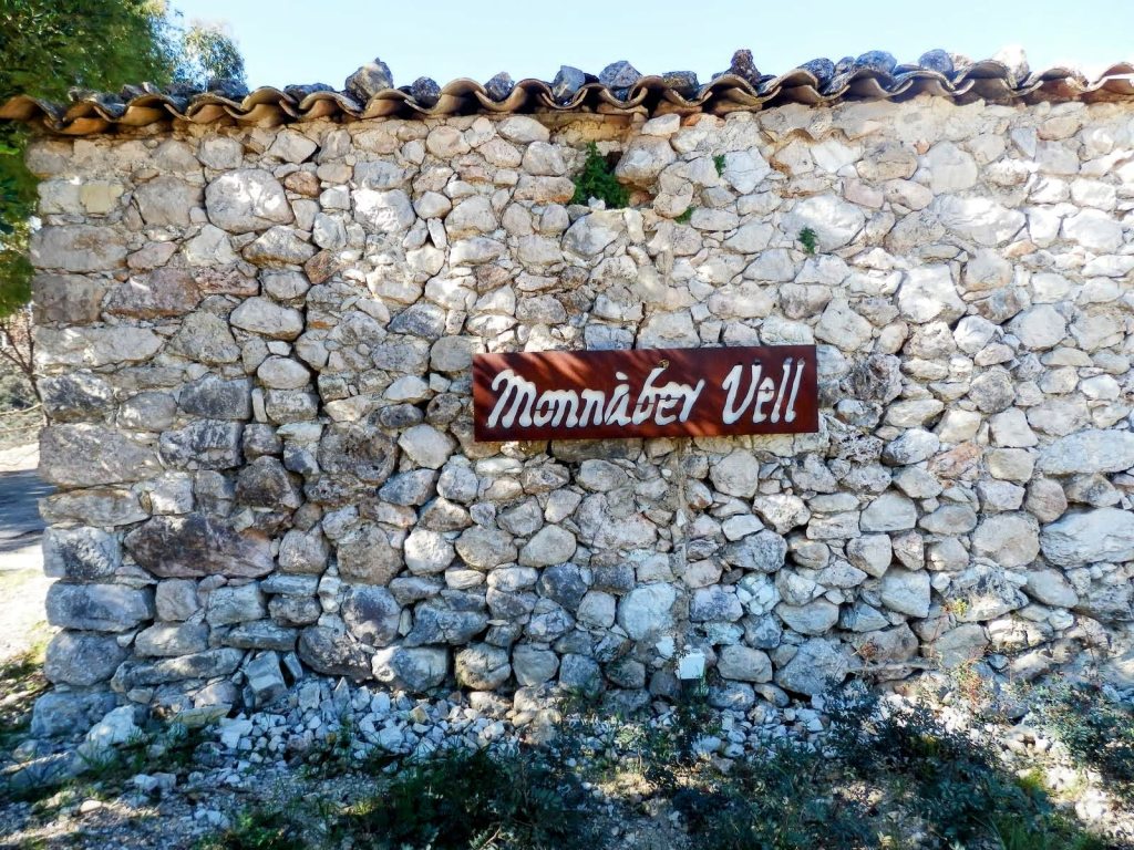 Pared de piedra seca con una placa de metal que dice 'Monnàber Vell' en un entorno natural.