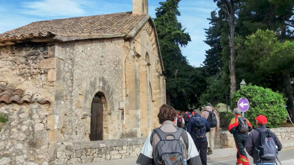 Grupo de personas caminando cerca de una iglesia de piedra con un tejado de tejas, rodeada de vegetación.