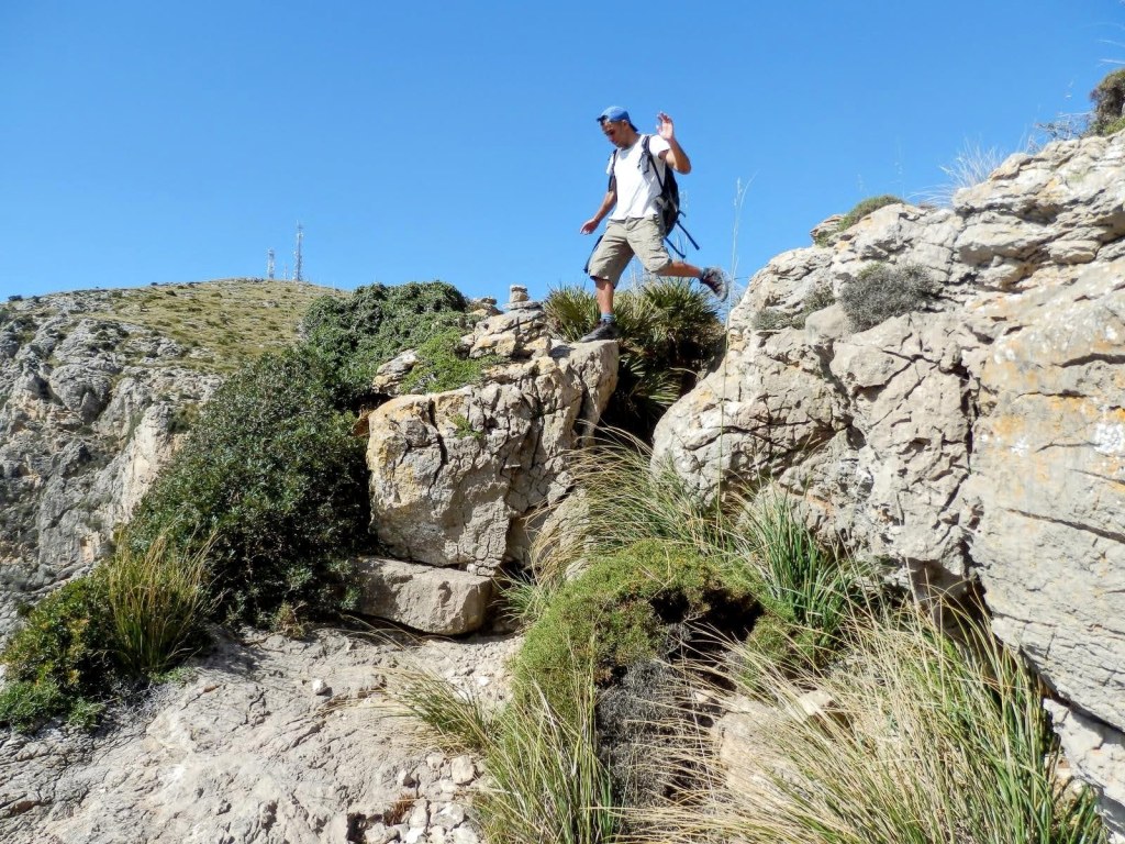 Senderista cruzando el Pas des Porcs cubierto de vegetación en una ruta montañosa, con un paisaje de colinas y un cielo despejado al fondo.
