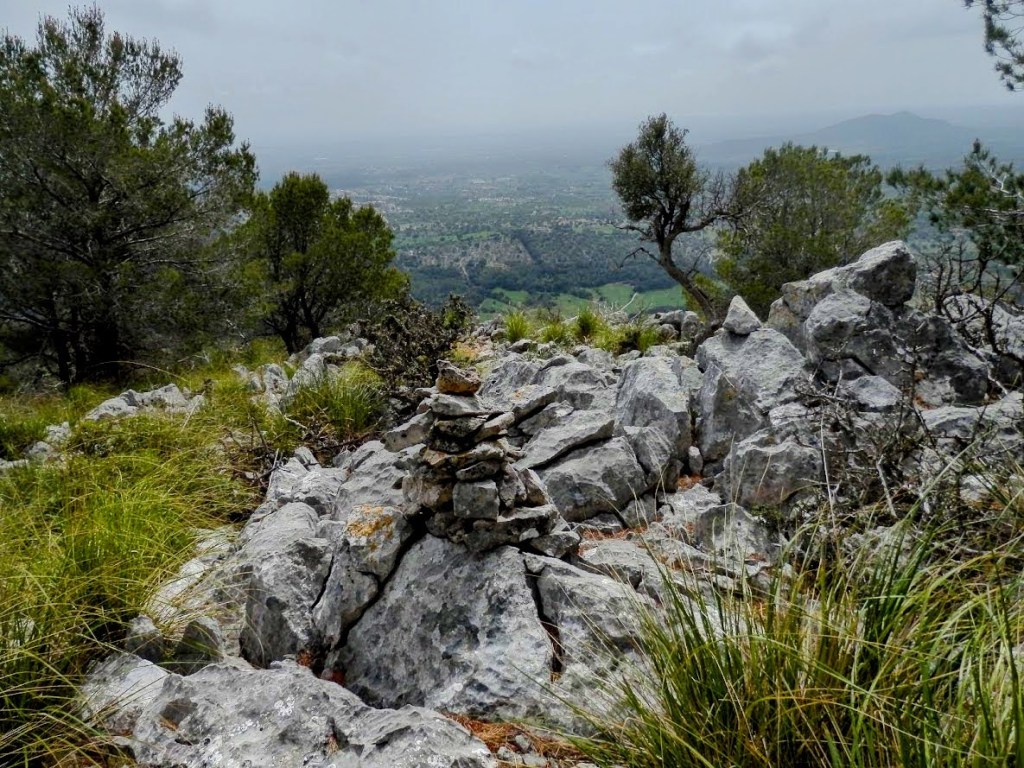 Vista panorámica desde la cima del Puig des Castell, con un hito de piedras en primer plano y paisajes verdes al fondo.