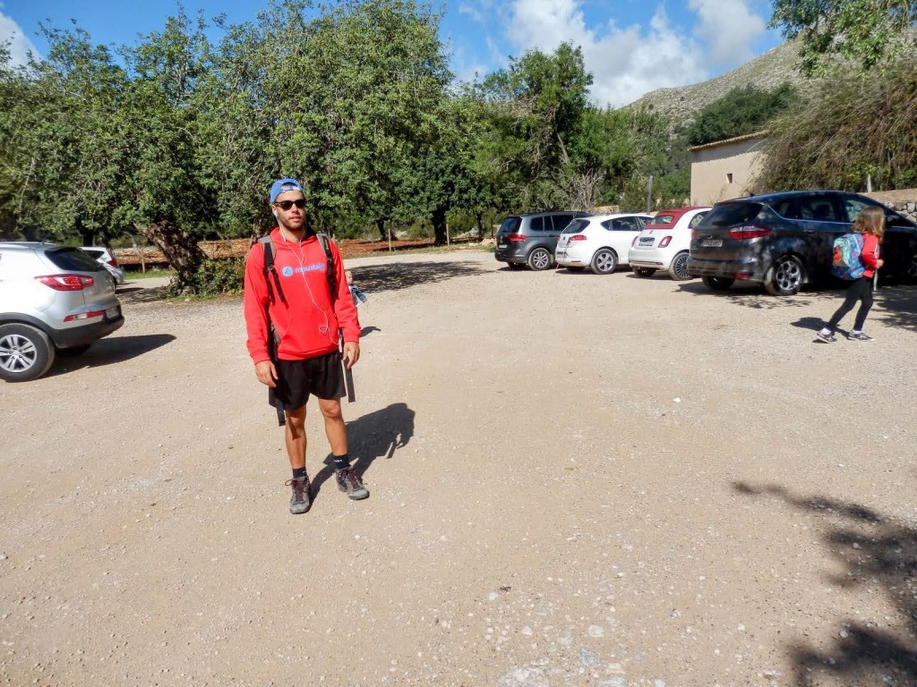 Hombre de pie en el aparcamiento de tierra de la casa de campo de l’amo en Biel, vistiendo una sudadera roja y pantalones cortos, con un paisaje de árboles y coches aparcados al fondo.