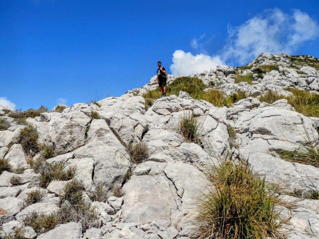 Un senderista ascendiendo por un terreno rocoso en el Puig de Galatzó, con un cielo azul y algunas nubes en el fondo.