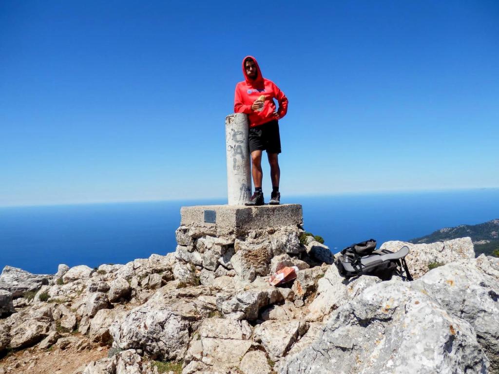 Persona de pie en la cima del Puig de Galatzó, sosteniendo un objeto, con el mar al fondo y un cielo despejado.