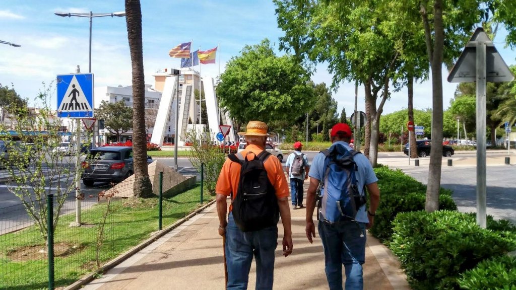 Grupo de personas caminando por un sendero junto a un área urbana, con señales de tráfico y árboles alrededor, con una estructura o monumento en el fondo.