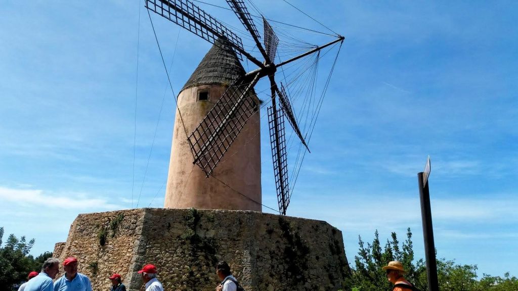 Molino de viento de piedra con aspas, rodeado de un grupo de personas en una jornada de senderismo bajo un cielo despejado.