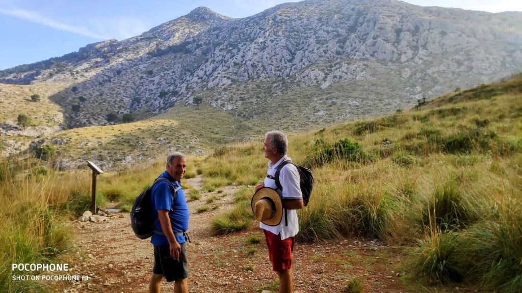 Dos senderistas charlando en un camino montañoso bajo un cielo despejado y rodeados de vegetación, con el Puig de Galatzó al fondo.