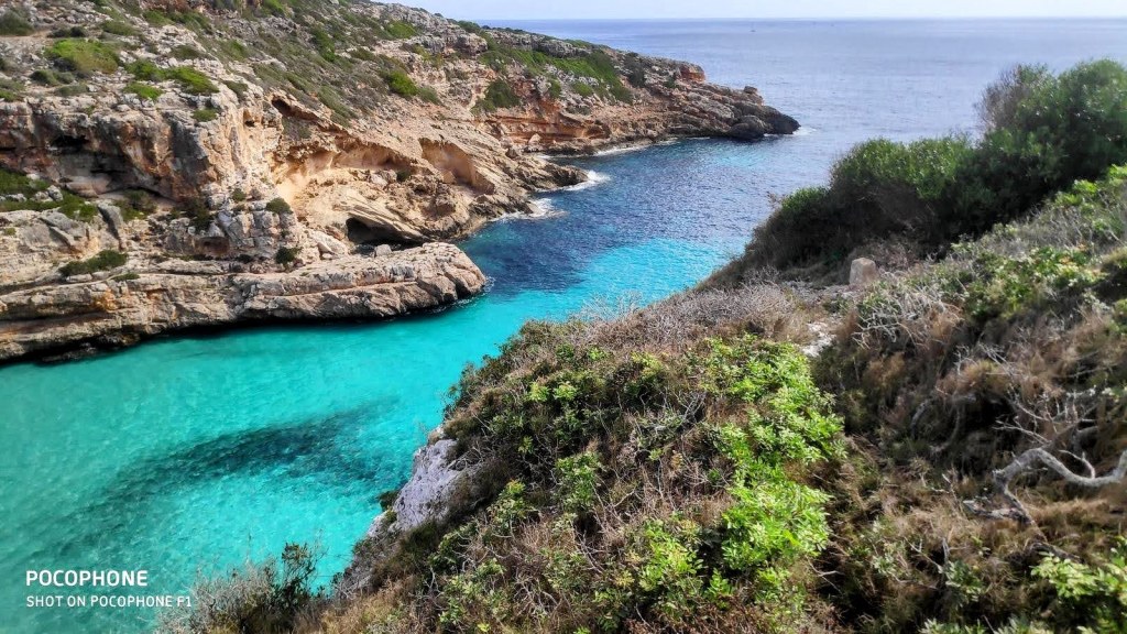 Vista de la costa cerca de Cala Marmols, con aguas turquesas y acantilados rocosos al fondo, rodeados de vegetación.