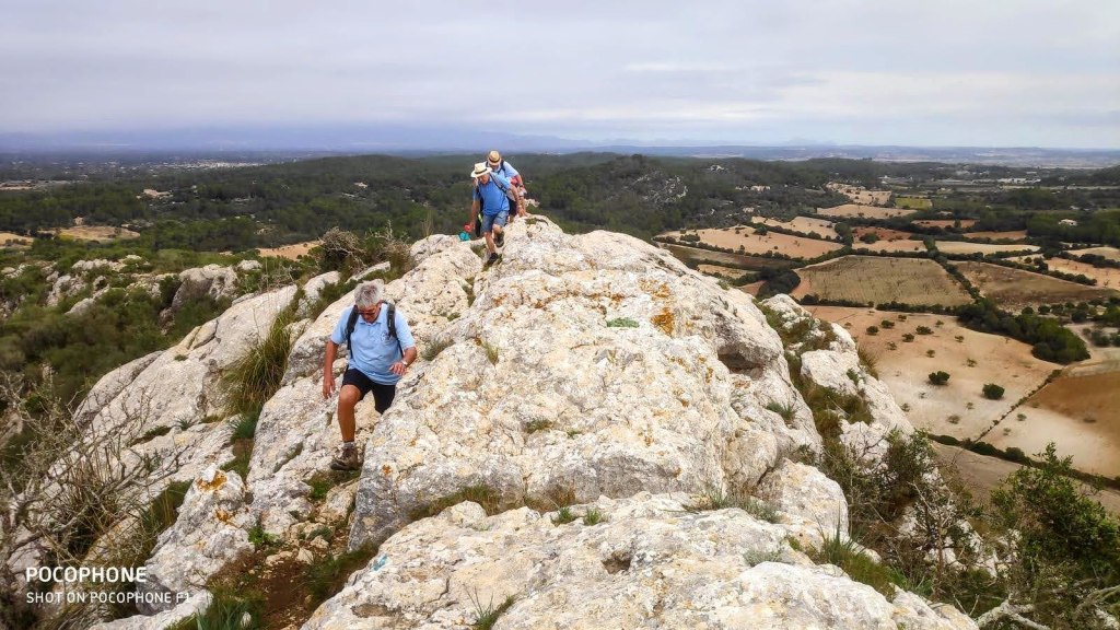 Grupo de personas caminando sobre una roca en la cima del Puig de ses Bruixes, con vistas panorámicas del paisaje de Llucmajor.