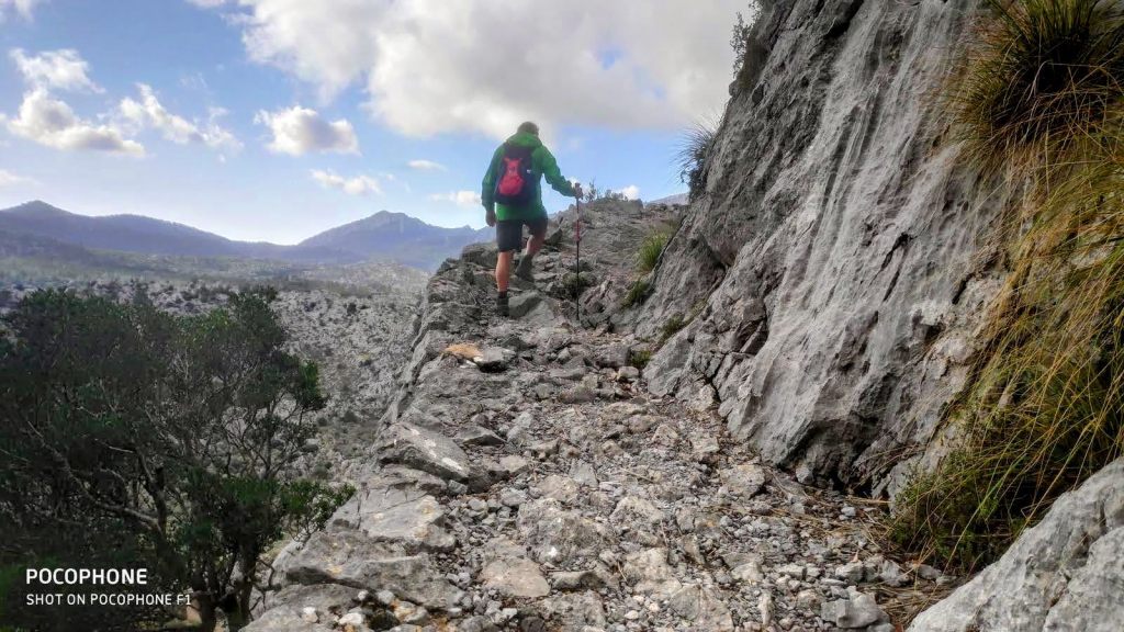 Persona caminando por Pas de ses Cases en la Sierra de Tramontana, con vistas a un paisaje rocoso y montañoso bajo un cielo parcialmente nublado.