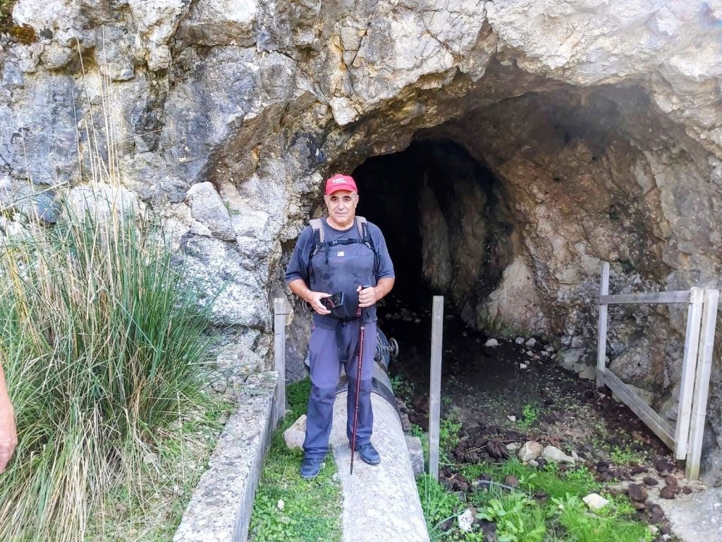 Una persona de pie frente a la entrada de un túnel en una zona rocosa, rodeada de vegetación con un bastón de trekking en la mano.