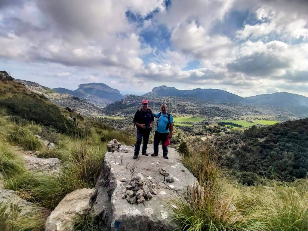 Dos personas de pie sobre una roca en un paisaje montañoso, con valles y montañas al fondo bajo un cielo nublado.