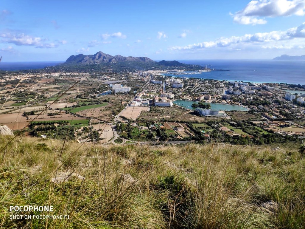Vista panorámica desde la cima del Puig de Sant Martí, mostrando la bahía de Alcudia y la ciudad de Puerto Alcudia, con montañas y campos verdes en el horizonte.