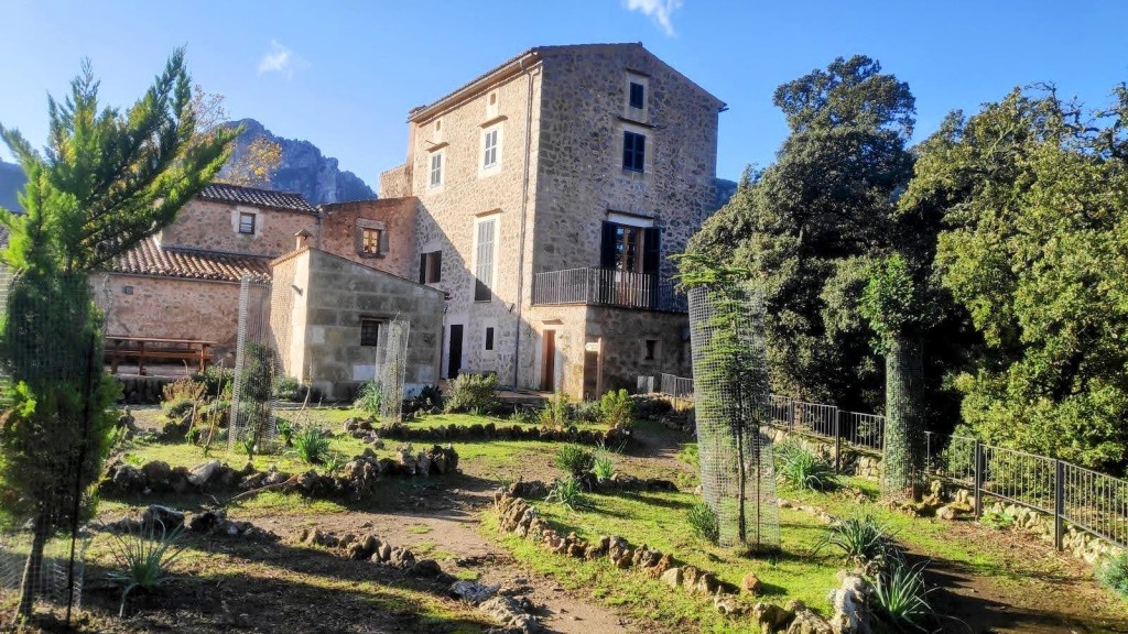 Vista del Refugio de Son Amer en medio de un jardín con vegetación y árboles, situada en un entorno montañoso.
