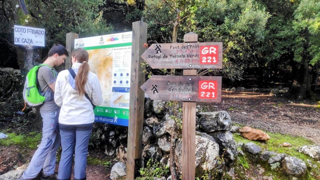 Dos personas observando un mapa en un panel informativo sobre rutas en la Serra de Tramontana, Mallorca, con señales que indican direcciones y tiempos de recorrido.