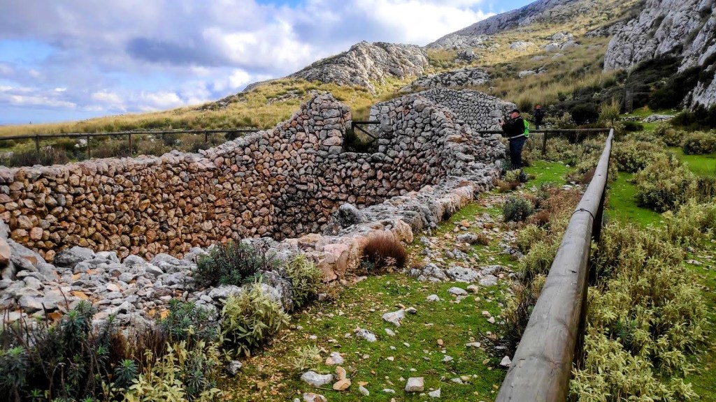 Vista de la Casa de Neu d’en Galileu, una estructura de piedra en ruinas rodeada de vegetación en la Ruta Puig d'en Galileu, con montañas al fondo.