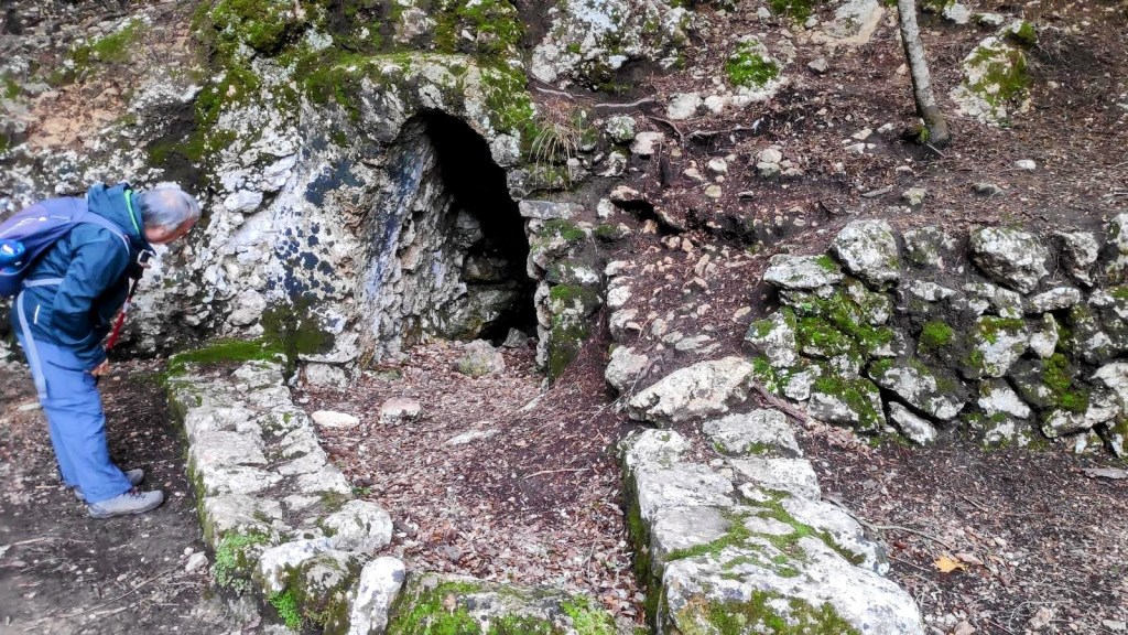 Persona examinando la entrada de la Fuente des Freu en un entorno forestal, rodeada de musgo y vegetación.