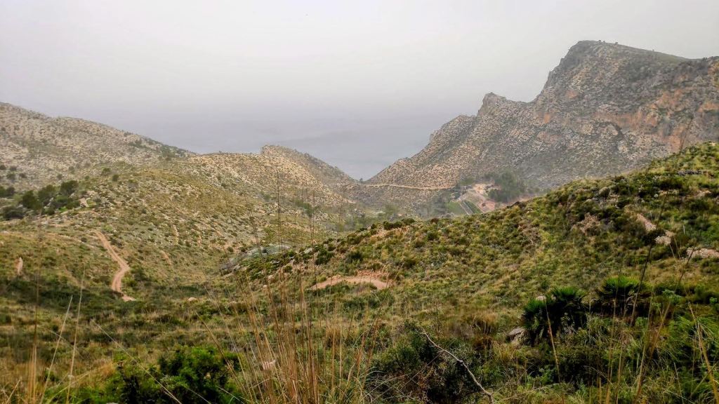 Vista panorámica de un paisaje montañoso con senderos y vegetación, mostrando la costa y las montañas de la zona de la Trapa en Mallorca, bajo un cielo nublado.