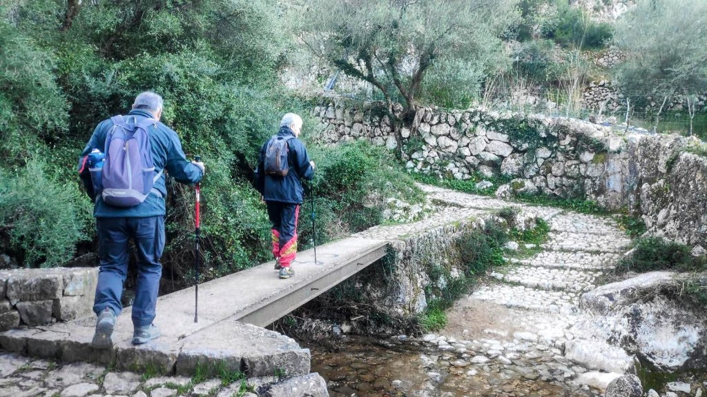 Senderistas cruzando un puente de piedra en el Barranco de Biniaraix, rodeados de vegetación y piedras.