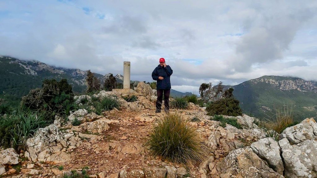 Una persona de pie en la cima del Puig de Sa Gubia, con un paisaje montañoso y nubes de fondo.