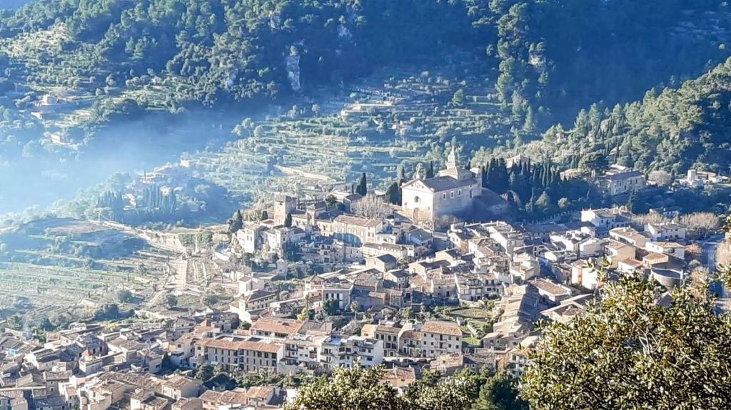 Vista panorámica de la localidad de Valldemossa, rodeada de montañas y vegetación, con edificios tradicionales y un ambiente tranquilo.