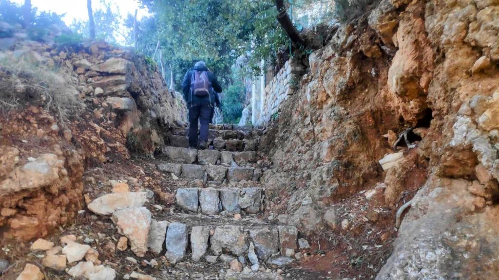 Persona subiendo por un camino empedrado rodeado de rocas y vegetación, en un entorno natural.