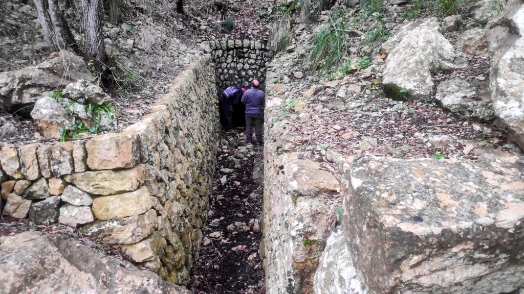 Vista de la Font des Puig con dos personas observando la estructura de piedra en un entorno natural.