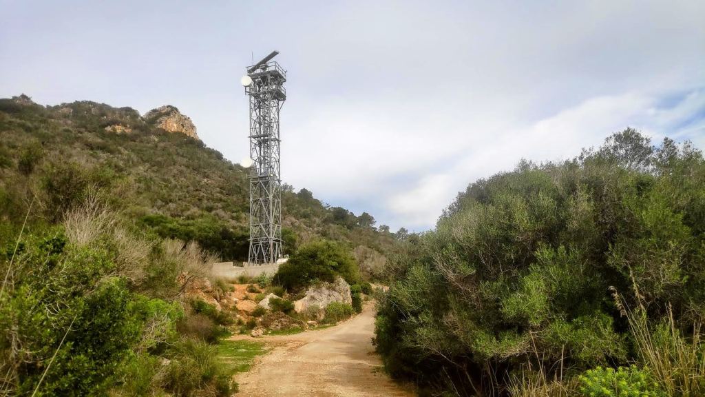 Vista de un camino rural rodeado de vegetación, con una torre metálica de telecomunicaciones al fondo, situada en un paisaje montañoso.