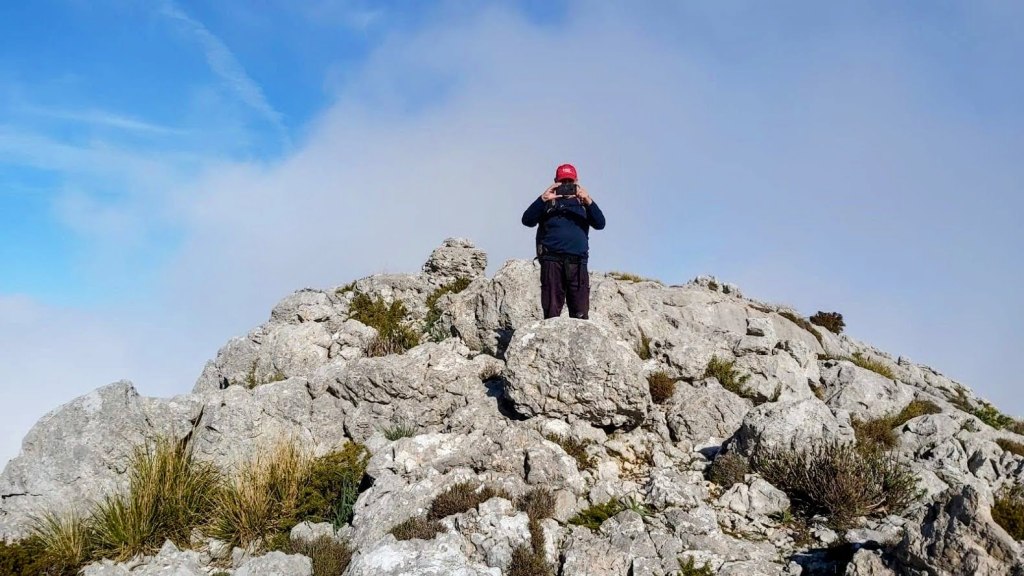 Un senderista en la cima rocosa del Puig de ses Vinyes, rodeado de nubes y un cielo azul, sosteniendo una cámara para tomar fotos.