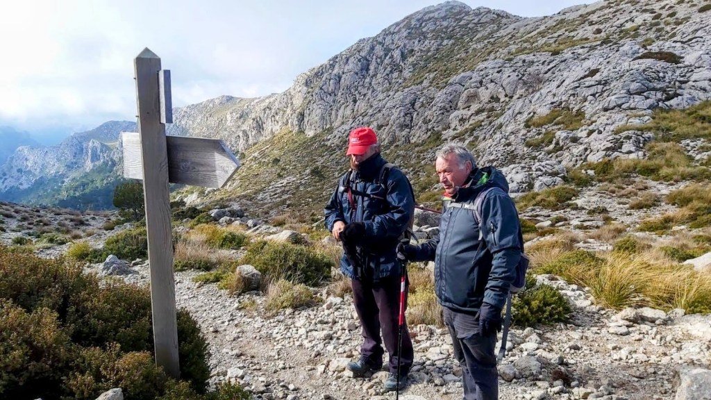 Dos senderistas en el Coll des Prat, observando un cartel de señalización en un paisaje rocoso y montañoso.